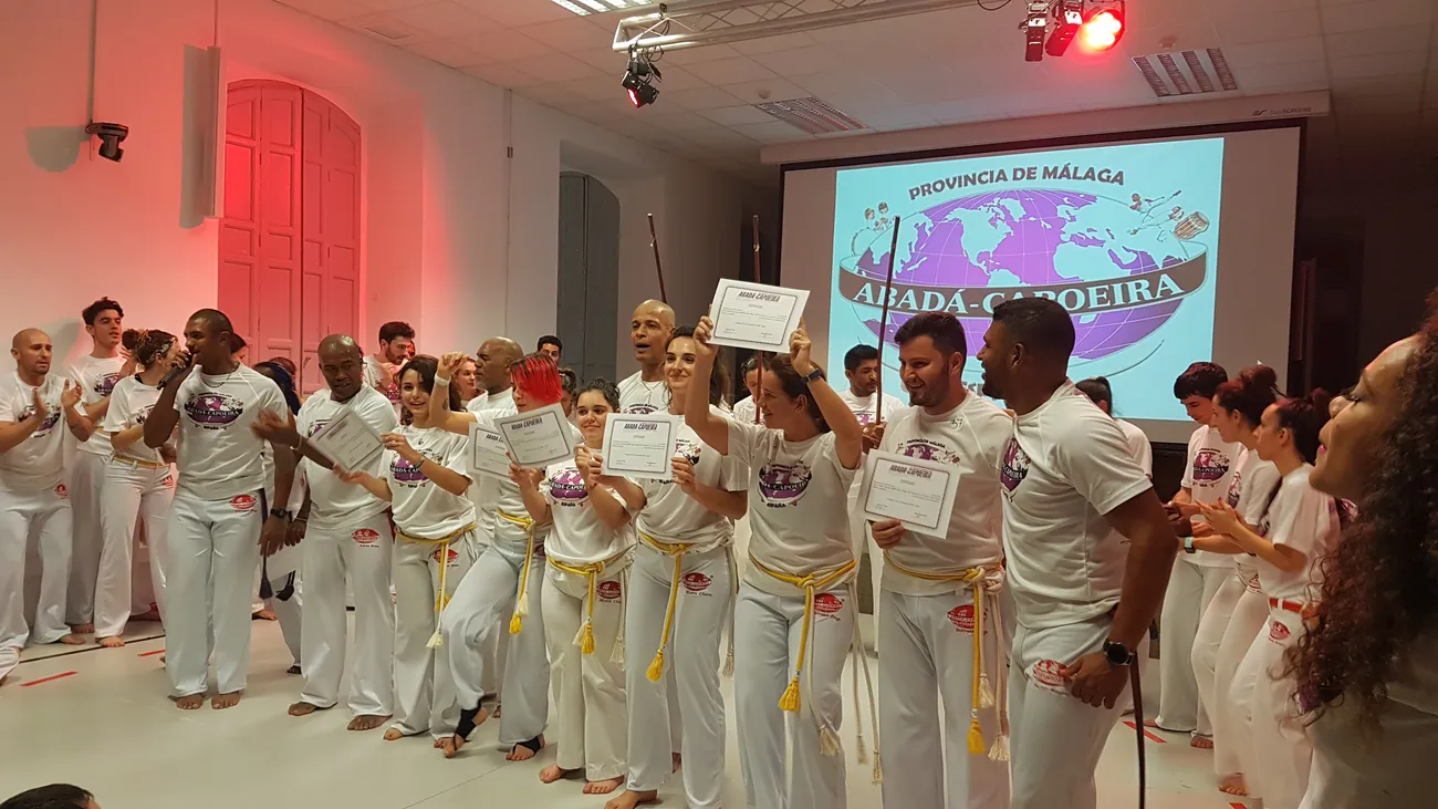 Alumnos de la escuela levantando sus diplomas al final de un batizado, con el logotipo de ABADÁ-Capoeira Provincia de Málaga proyectado detrás