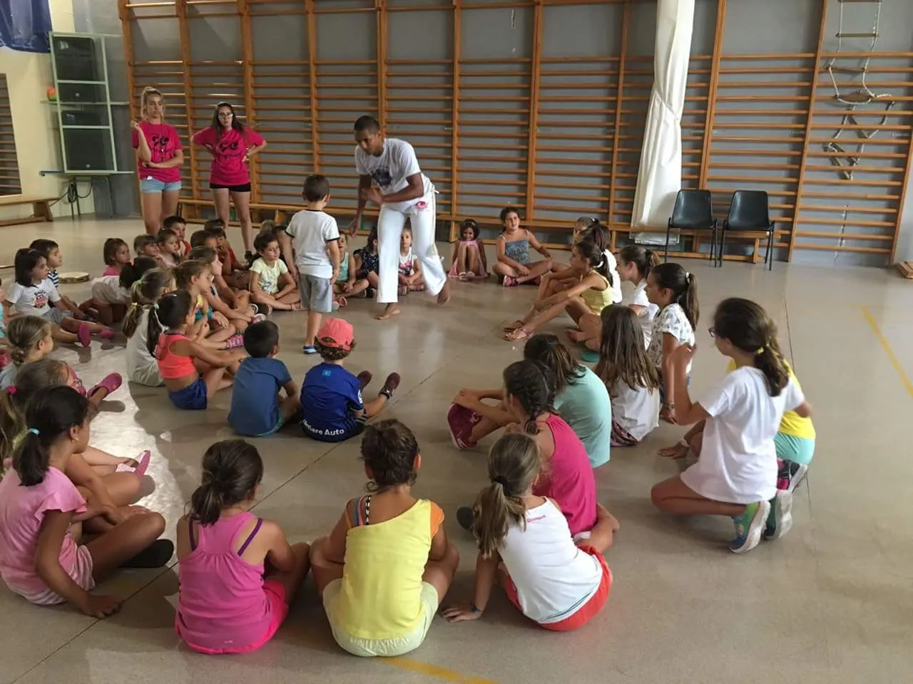 Niños y niñas sentados en círculo en un pabellón durante un taller de capoeira