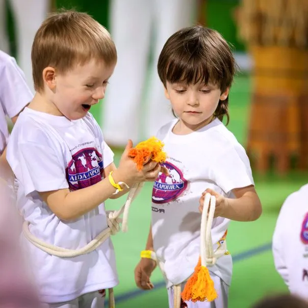 Dos niños pequeños con camiseta de ABADÁ-Capoeira sujetando una corda con borlas naranjas